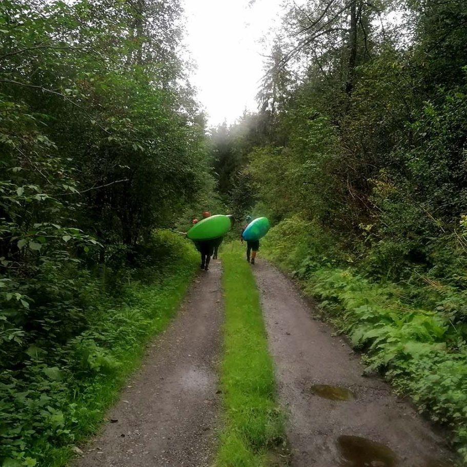 Zwei Kajakfahrer tragen grüne Kajaks auf einem schmalen, bewaldeten Weg.