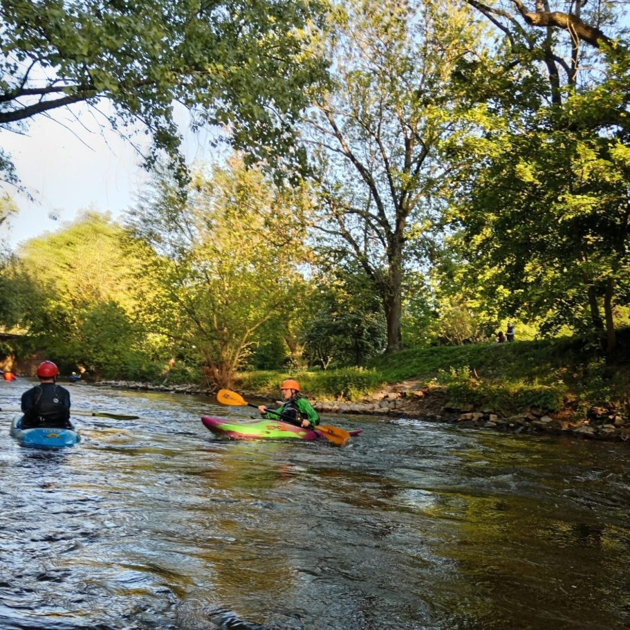 Jugendtraining Zwei Kajakfahrer paddeln auf einem ruhigen Fluss, umgeben von Bäumen und Sonne.