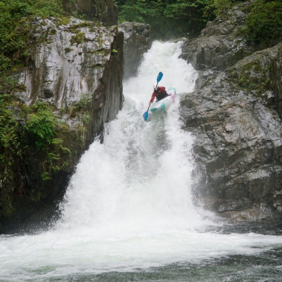 Kajakfahrer stürzt einen Wasserfall hinunter, umgeben von Felsen und grüner Vegetation.