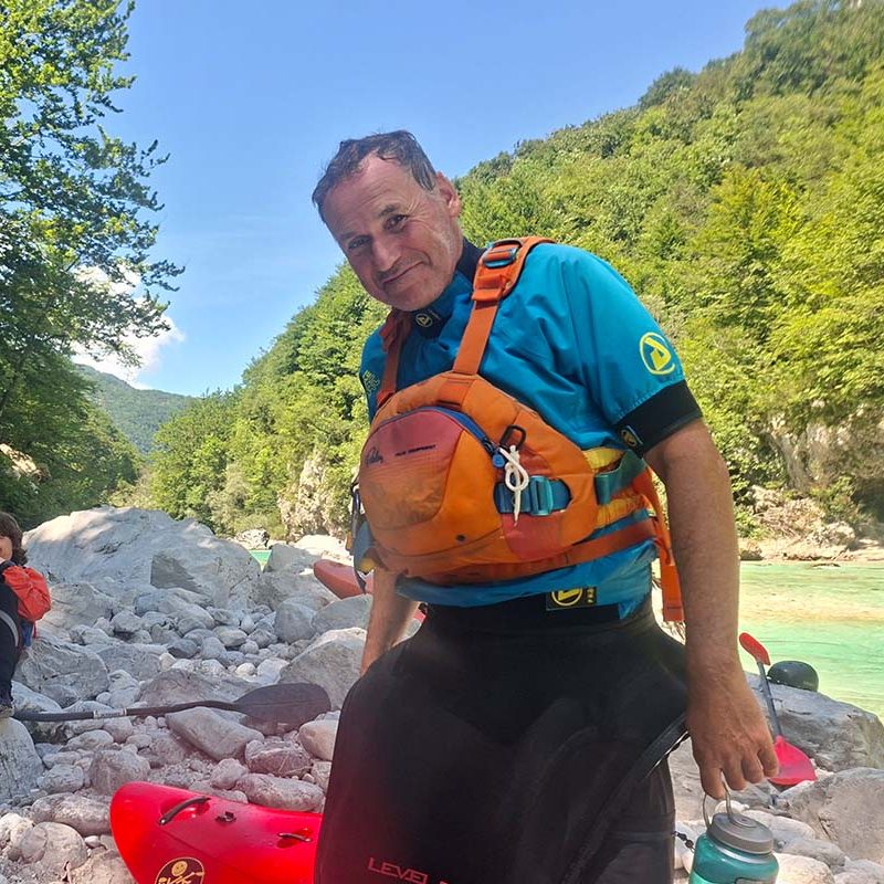 Mann in einem blauen Shirt und Rettungsweste steht am Ufer eines Flusses.