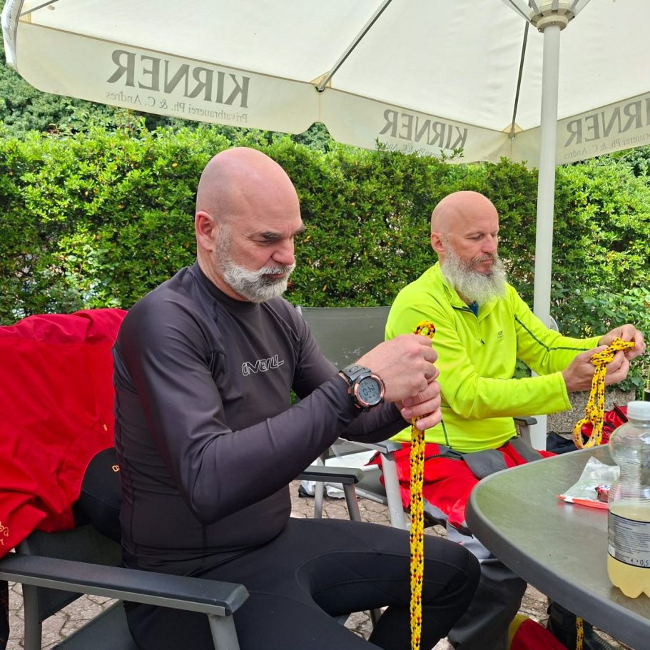 Sicherheitslehrgang: Übung Achterknoten Zwei Männer in Outdoor-Bekleidung knoten Seile am Tisch unter einem Sonnenschirm.