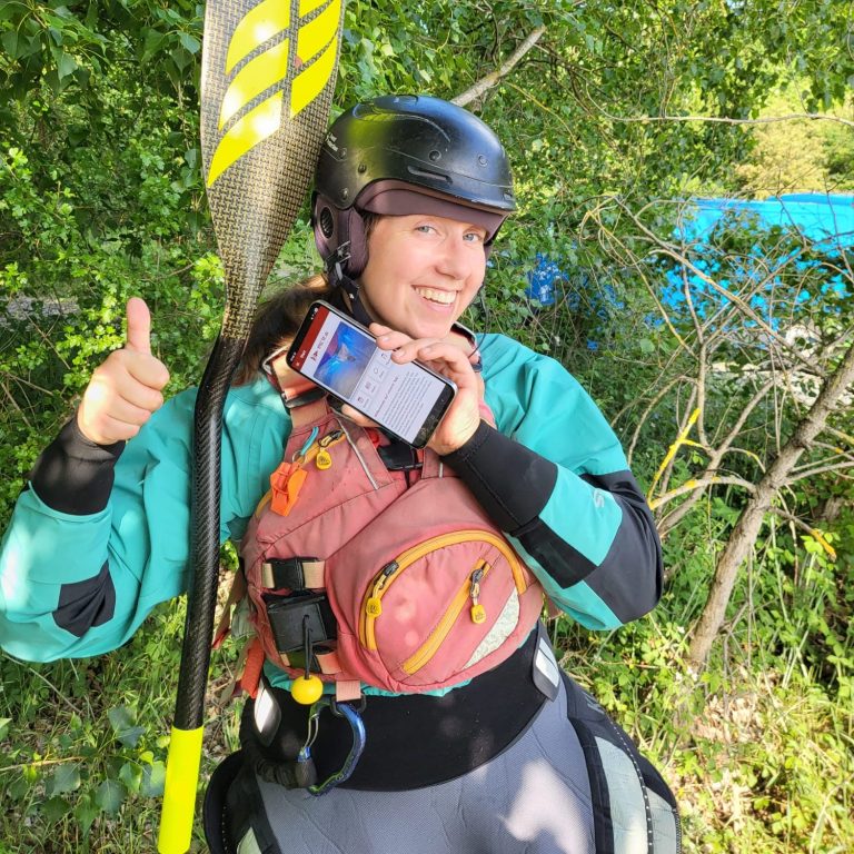 Person hält ein Smartphone am Fluss, mit der HKCW-Vereinsapp-Anzeige auf dem Bildschirm.