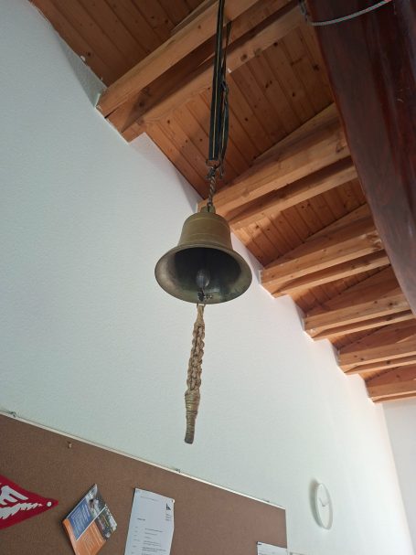 Vereinsglocke Eine hängende Glocke mit einer Schnur an einer holzverkleideten Decke.