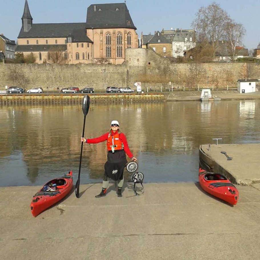 Person mit Paddel steht an einem Steg zwischen zwei roten Kajaks.