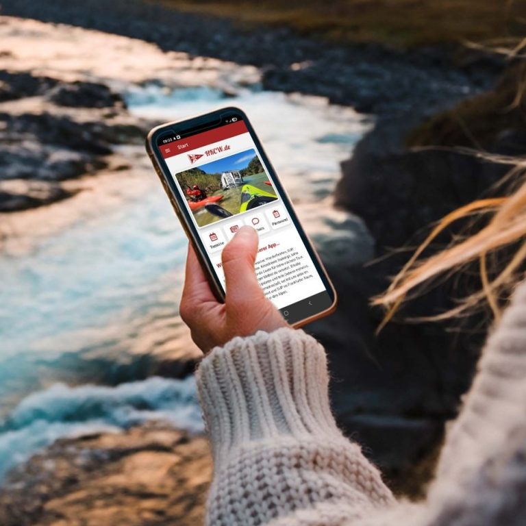 Person hält ein Smartphone am Fluss, mit der HKCW-Vereinsapp-Anzeige auf dem Bildschirm.