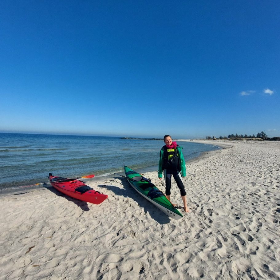 Seekajaktour Person steht am Strand neben zwei Seekajaks bei klarem, sonnigem Wetter.