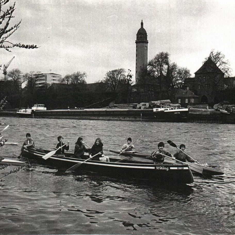Ruderer in einem Boot auf einem Fluss, im Hintergrund das Höchster Schloss und Bäume.