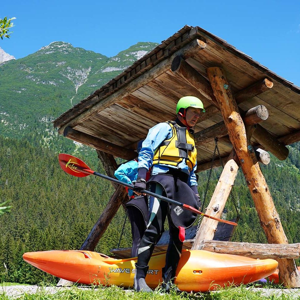 Paddler mit Kajak und Paddel vor einer Holzhütte in Berglandschaft.