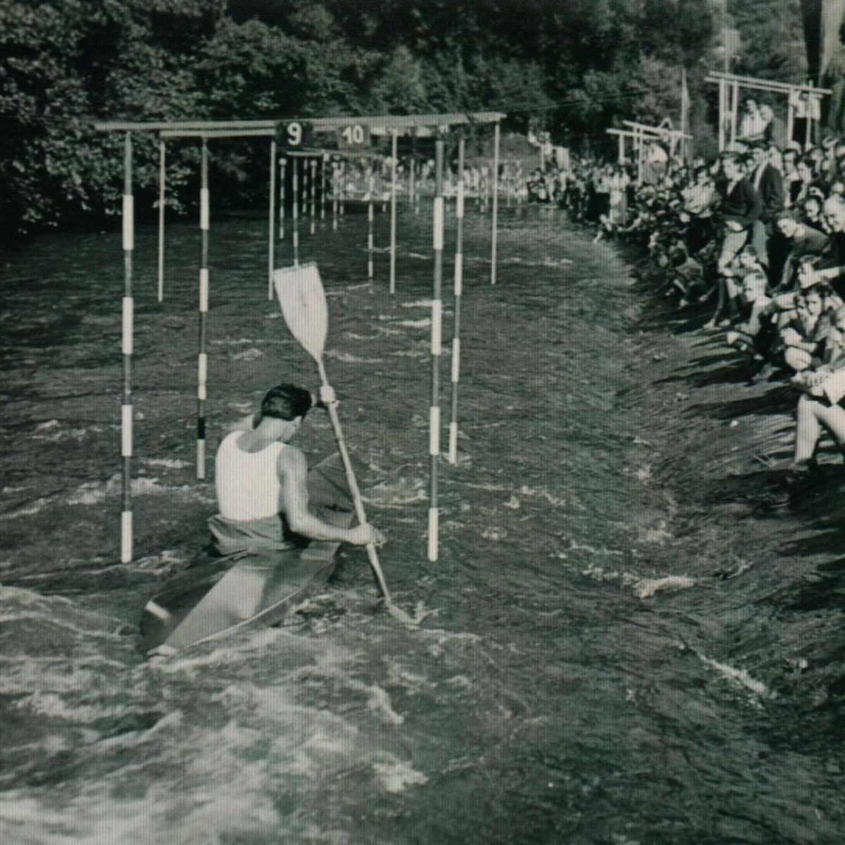 Ein Kanute paddelt durch ein Slalom-Parcours, umgeben von Zuschauern am Ufer.