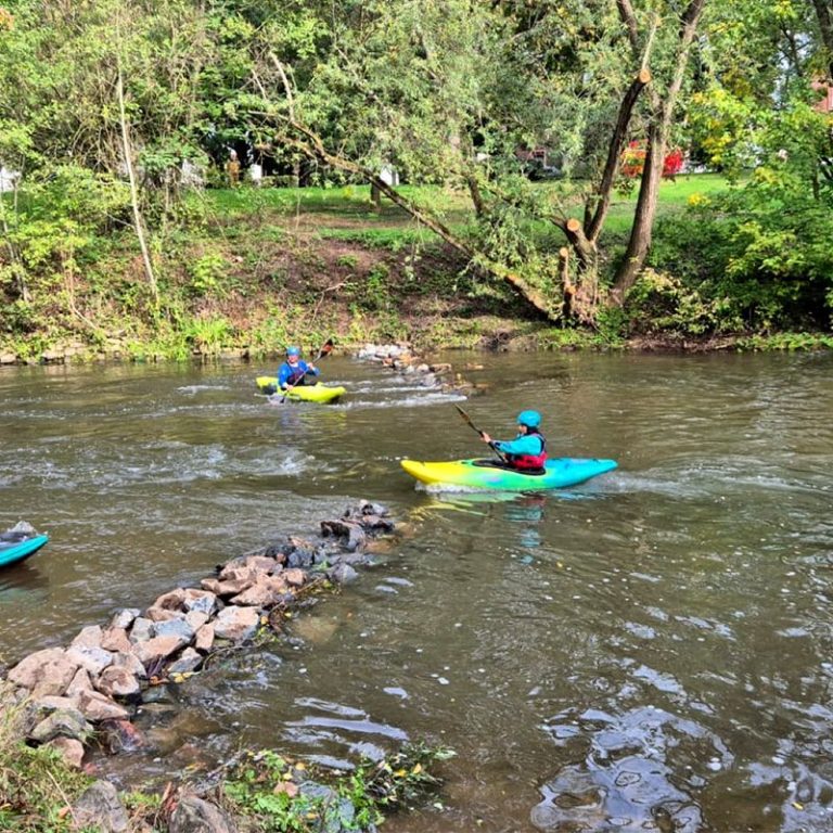 Zwei Personen in Neoprenanzügen mit Schwimmwesten, lächelnd an einem Ufer.