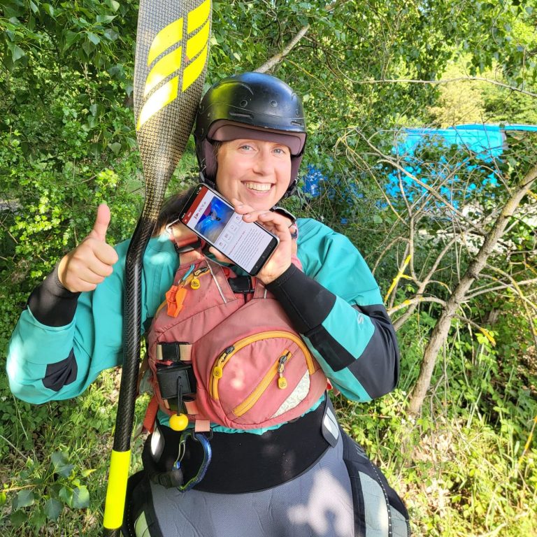 Person hält ein Smartphone am Fluss, mit der HKCW-Vereinsapp-Anzeige auf dem Bildschirm.