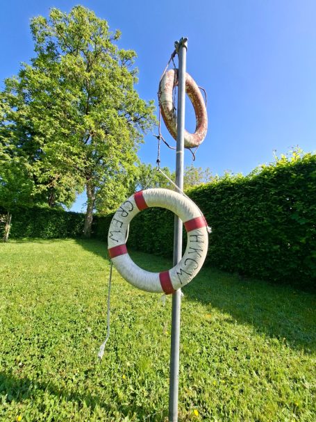 Rettungsring mit der Aufschrift "HKCW-Crew" Rettungsringe hängen an einem Pfosten vor einer grünen Hecke unter blauem Himmel.