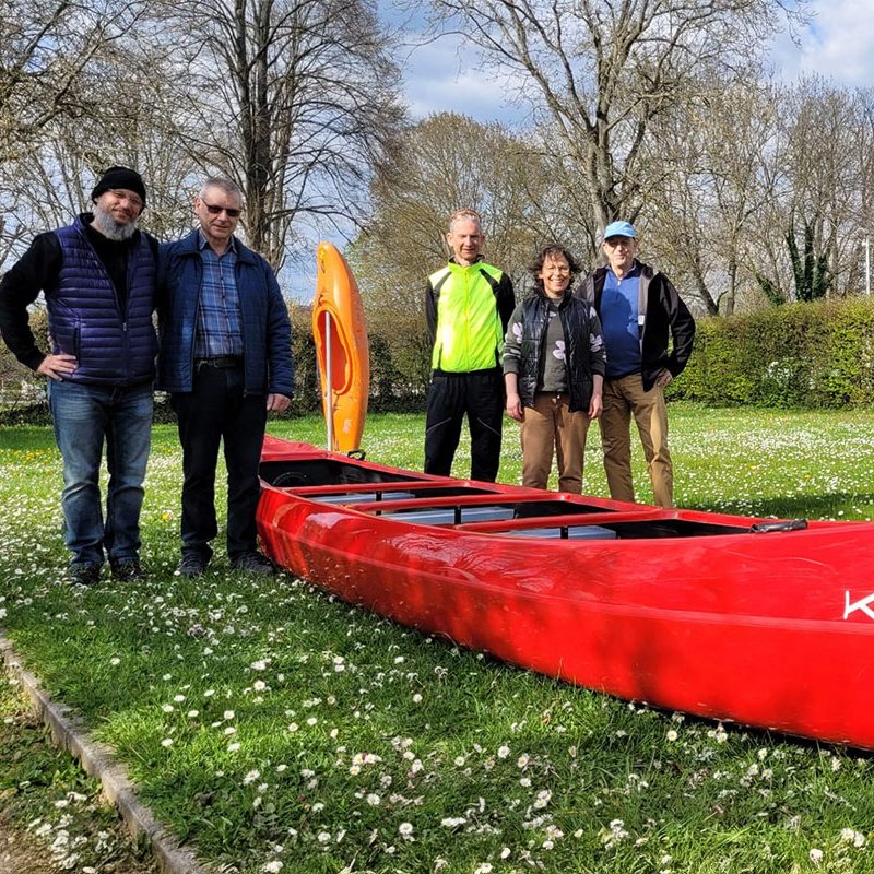 Gruppe von fünf Personen neben einem roten Kajak auf einer Wiese mit Blumen.