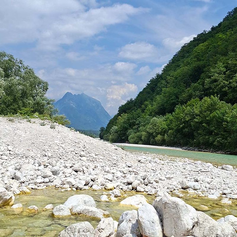 Ein klarer Fluss, umgeben von grünen Bergen und Kiesstränden unter einem blauen Himmel.